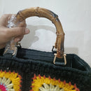 A close-up of a crocheted handbag with a curved bamboo handle, held by a hand. The bag has a black crochet body with yellow, pink, and white granny square patterns.