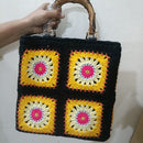A handmade crocheted tote bag with a black base and four yellow, white, and pink granny square patterns, featuring a natural bamboo handle held by a hand against a plain white wall.