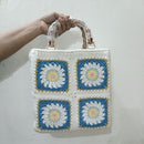 A handmade crocheted handbag with a white base and four blue floral granny square panels, held by a hand wrapped in plastic against a plain white wall.